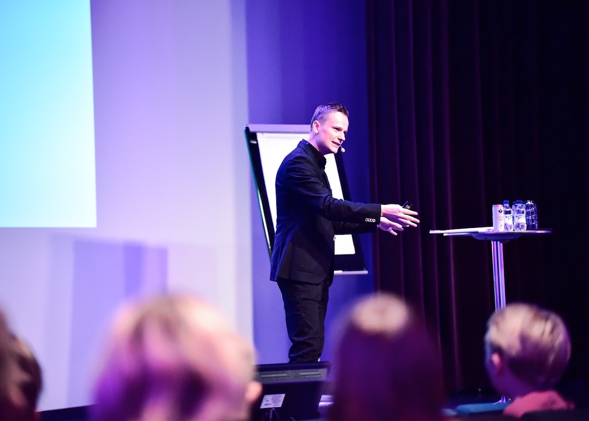 Henri pitää esitystä ja puhuu mikrofonin kanssa lavalla yleisön edessä.