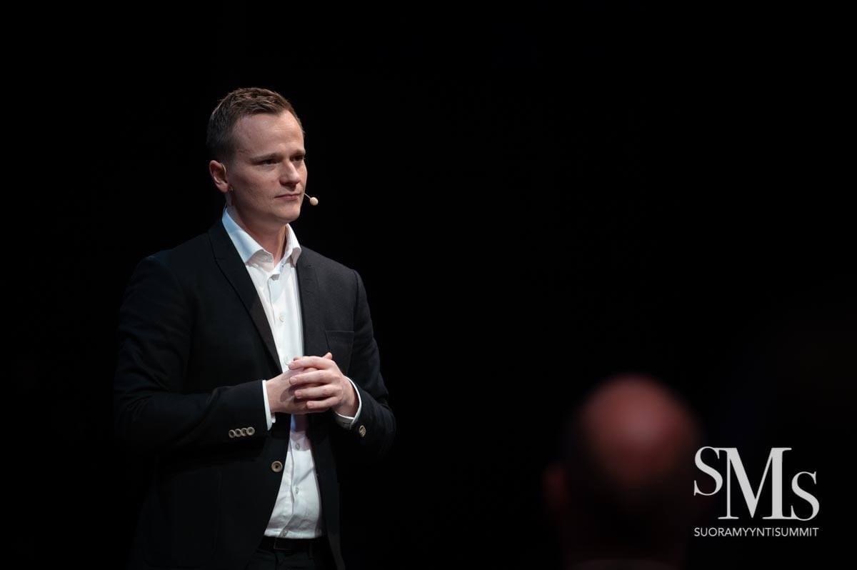 Henri puhuu mikrofonikuulokkeella mustassa puvussa valkoisen paidan kanssa.